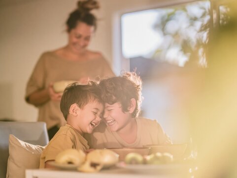 Two young brothers laughing together at a kitchen table while their mother watches fondly during a sunlit morning breakfast for family lifestyle storytelling