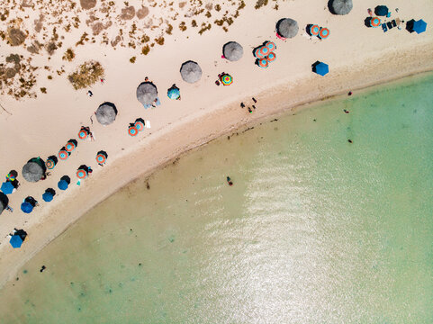 Aerial view of Balandra beach with colorful umbrellas lining the sandy shore and people swimming in the clear turquoise water of La Paz, Baja California Sur, Mexico.