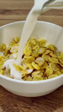 Fresh milk pouring into a white bowl of crispy corn flakes. Healthy and delicious morning meal preparation on a wooden kitchen table