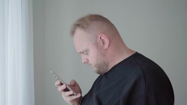 Middle aged man hunches over his device by a bright window, showing unhealthy neck posture from extended screen use.
