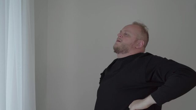 Middle aged man in a black top stretches his stiff back and side muscles in a plain indoor room for pain relief.
