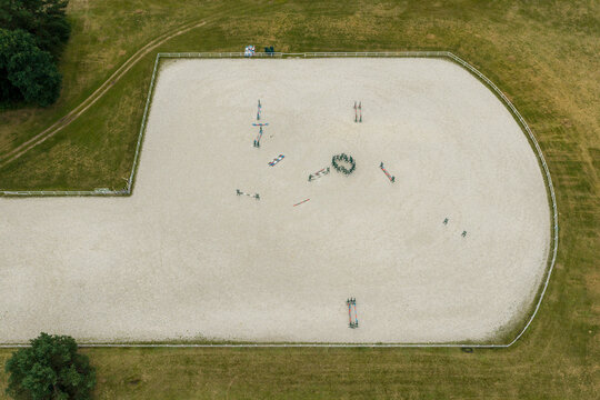 Aerial view of an equestrian training arena with sand surface, scattered jumps, and a group of people standing in a circle in the center, Anyksciai, Utena County, Lithuania.