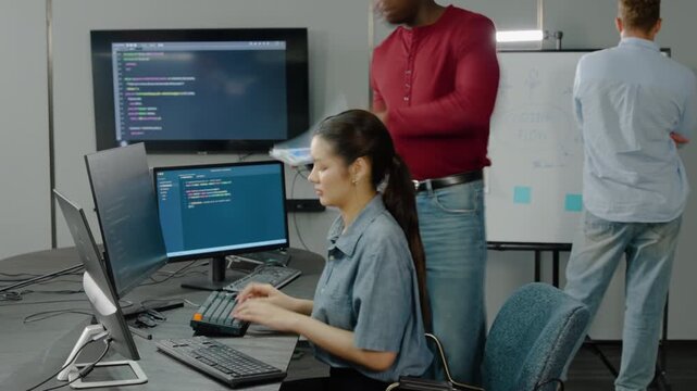 Diverse software development team working fast in a modern tech office. Asian female programmer typing code on computer while male colleagues collaborate on whiteboard in this agile startup workspace.
