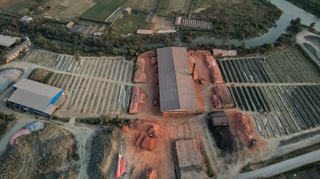 Aerial view of a brick kiln facility with rows of drying bricks, a large production building, and surrounding agricultural fields near a river in Chittagong, Chattogram, Bangladesh.