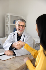 Fototapeta premium Smiling senior male doctor shaking hands with female seated at desk in clinic office, represents professional medical consultation services, qualified patient care, and trust in healthcare industry