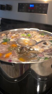 Large stainless steel pot with homemade soup boiling on a kitchen stove, filled with colorful vegetables, mushrooms and meat. Close-up view of a hearty meal cooking 