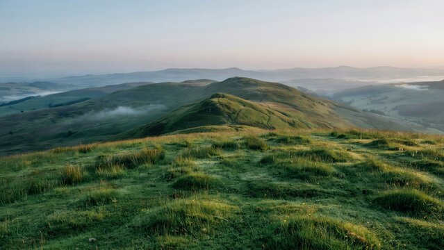 Rolling green hills covered in lush grass under a soft morning sky with patches of mist settling in the valleys of a peaceful rural landscape