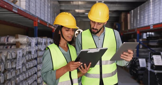 of young Hispanic warehouse workers in hardhats checking electronic and paper invoices together, comparing data on tablet and in file, working on inventory, standing at storage metal shelves