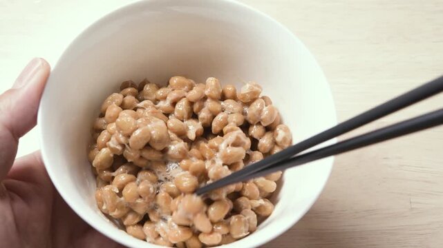 Chopsticks stirring fermented natto beans until sticky and stringy in white bowl, static shot
