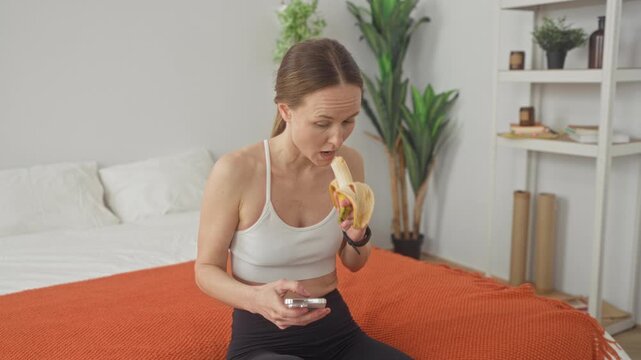 Woman eating a peeled banana while checking a smartphone on a bed in a studio; post workout morning calm.