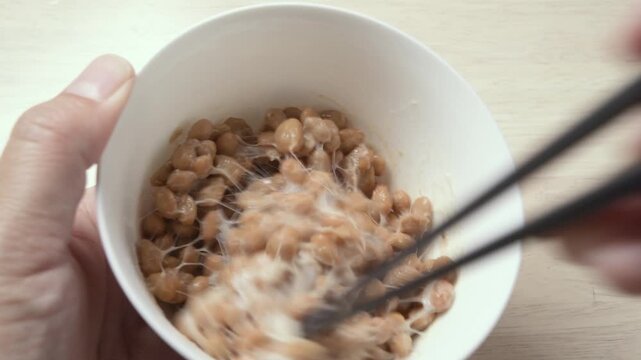 Chopsticks stirring fermented natto beans until sticky and stringy in white bowl, static shot