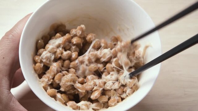 Chopsticks stirring fermented natto beans until sticky and stringy in white bowl, static shot