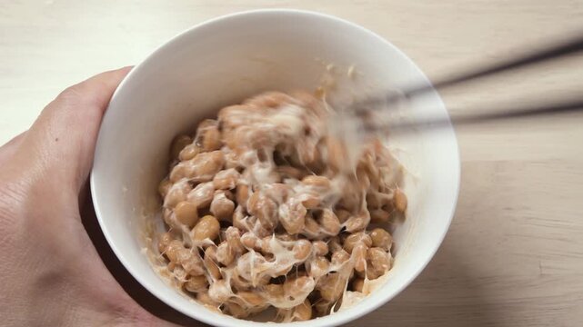 Chopsticks stirring fermented natto beans until sticky and stringy in white bowl, static shot