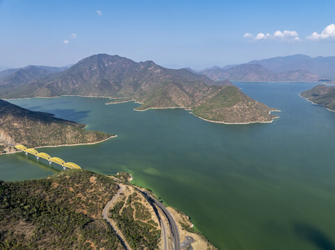 Aerial view of the yellow bridge spanning the Rio Balsas reservoir, surrounded by rugged mountains and green water under a clear blue sky in Infiernillo, Michoacan, Mexico.