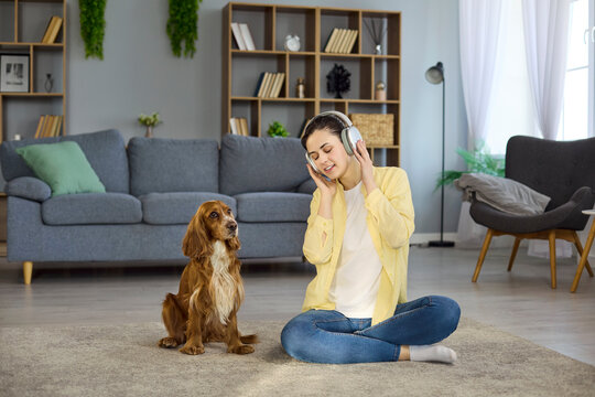 Woman listening to music with headphones, spaniel dog. At home the owner sits on the carpet, smiling as the attentive pet dog watches, capturing a cozy moment. Clear concept of pet bonding.