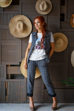 Caucasian redhead woman in a grey checkered suit beside a wall of straw hats in a rustic wooden interior, full height frame for millinery, retail and summer accessory content