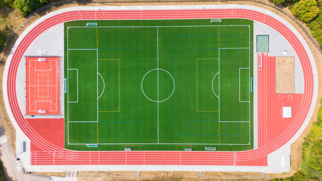 Aerial view of a sports ground featuring a green turf football field, a red running track, and a basketball court, surrounded by trees in Trier-Saarburg, Rhineland-Palatinate, Germany.