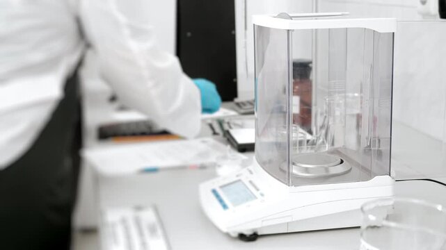 Laboratory technician cleans a precision analytical balance with a brush inside its protective chamber, maintaining scientific equipment for accurate measurements.