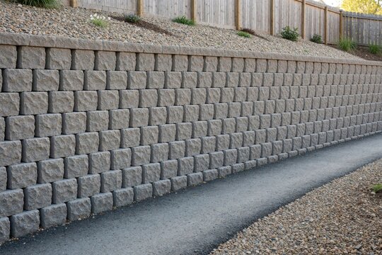 Stone block retaining wall supports sloped terrain next to paved path. Wall structure prevents soil erosion near wooden fence and gravel area.