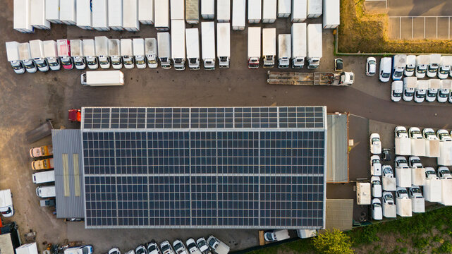 Aerial view of an industrial building with rooftop solar panels, surrounded by a parking lot filled with semi-trucks and construction machinery in Trier-Saarburg, Rhineland-Palatinate, Germany.