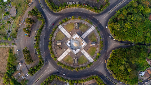 Aerial view of the Simpang Lima Gumul monument at the center of a large circular roundabout surrounded by lush palm trees and radial roads in Kediri, East Java, Indonesia.