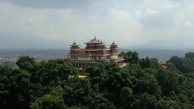 Elevated frontal aerial shot of a grand Buddhist monastery with layered gold roofs standing prominently above a green forested ridge in Nepal