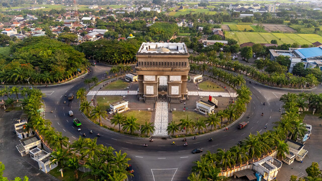 Aerial view of the Simpang Lima Gumul Monument, a grand archway in the center of a large roundabout surrounded by palm trees and urban infrastructure in Kediri, East Java, Indonesia.