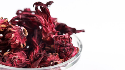 A close-up, macro composition highlighting dried dark red roselle hibiscus flowers in a glass bowl, set against a clean white backdrop with plenty of room for text on the right. © VH_World