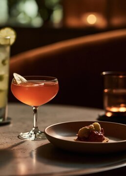Close-up of a cocktail glass on a wooden table. the glass is filled with a pinkish-red liquid and has a slice of lemon on the rim. next to the glass, there is a small plate with a dessert on it.