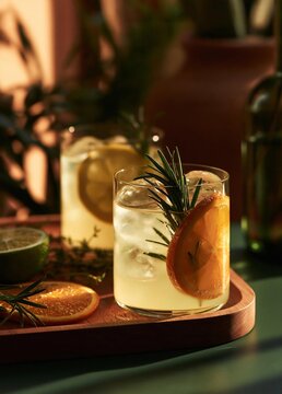 Two glasses of a refreshing drink on a wooden tray. the glasses are filled with ice cubes and have a slice of orange and a sprig of rosemary on the rim.