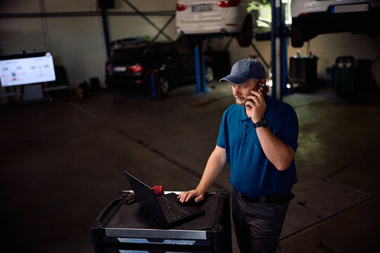 Car mechanic communicating on cell phone while working on laptop in repair shop.