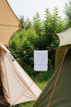 Two tents set up outdoors with a white towel drying on a clothesline between them. Green foliage and trees form the background. Summer camping scene.