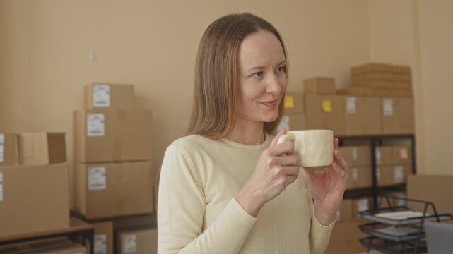 Young woman holding coffee mug and smiling while standing among stacked shipping boxes in building; small business contentment.