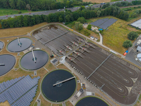 Aerial view of a wastewater treatment plant featuring circular sedimentation tanks and long aeration basins, with a large solar panel array and a highway in the background, Netherlands.