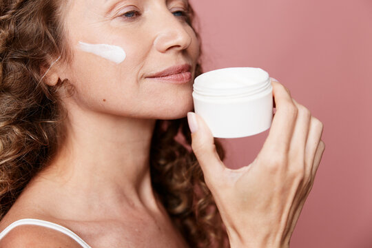 An authentic skincare moment with a middle aged woman applying cream to her cheek, smiling softly in a pink studio. Natural skin, no retouching, candid self care.
