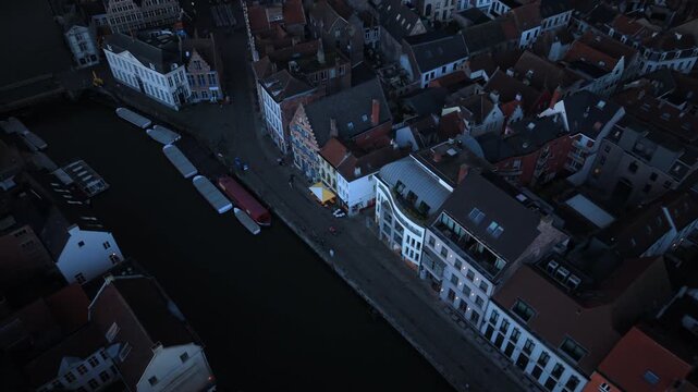 Cinematic aerial Ghent Belgium view capturing historic canal waterways with moored tour boats and traditional Flemish architecture. Red rooftops and gabled houses line the waterfront at twilight.