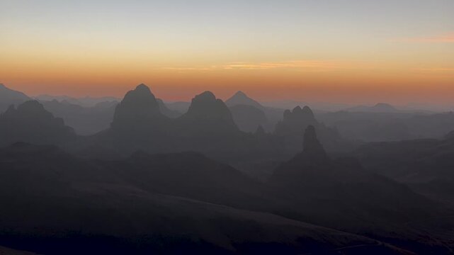 A breathtaking view of the dramatic, jagged peaks of the Hoggar Mountains in Algeria, silhouetted against a vibrant, colorful sunrise sky at dawn.
