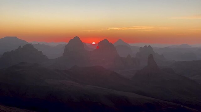 A stunning panoramic view of the sun rising behind the dramatic, jagged rock formations of the Hoggar Mountains in the Sahara Desert, Algeria.