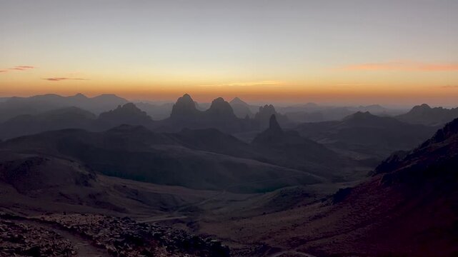 A stunning panoramic view of the dramatic volcanic peaks of the Hoggar Mountains in Algeria during a serene and golden sunrise, showcasing vast desert landscape.