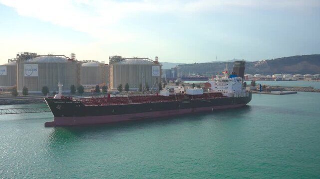 Large crude oil tanker ship maneuvering slowly in the calm waters of a major industrial sea port, with massive fuel and gas storage tanks visible on the coastline under a bright, sunny sky