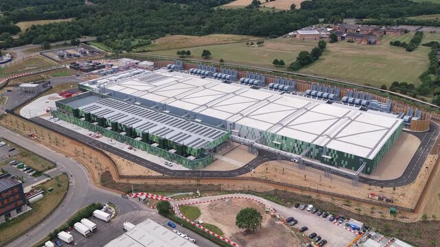 Large industrial buildings house servers in a rural area surrounded by green fields and parking spaces during a clear day as cars drive along the perimeter roads of the complex
