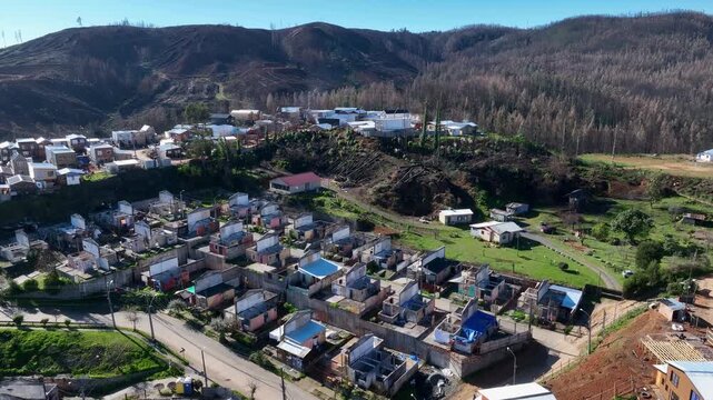 Circular flight around residential neighborhood with rebuilding rooftops and blue tarps surrounded by burned hillside and scattered pine trees. Lirquen, Penco, Chile