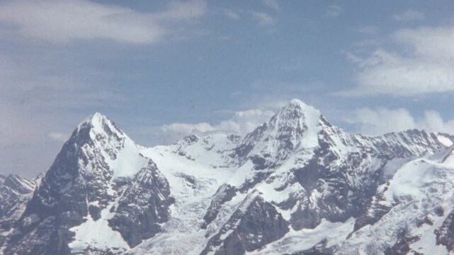 This archival footage from a film reel presents a static, wide shot of a majestic snow-capped mountain range in the Swiss Alps. The two prominent peaks dominating the frame are identifiable as the