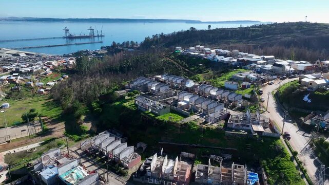 Circular flight around rebuilding neighborhood with new rooftops and construction frames revealing the industrial pier, calm bay and distant coastline. Lirquen, Penco, Chile
