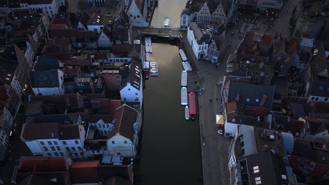 Cinematic aerial Ghent Belgium reveals medieval architecture lining waterways with moored tour boats. Historic gabled houses and bridges frame the canal as evening light bathes the Flemish cityscape.
