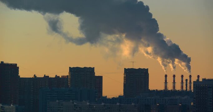 Dark factory smoke billows from a row of industrial chimneys behind silhouetted residential towers as low golden sunlight reveals dense layers of emissions spreading across the urban sky at dusk