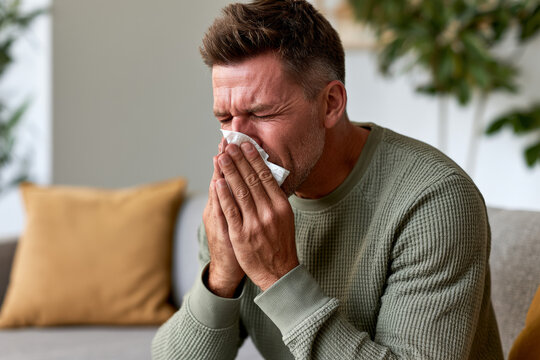Adult man sneezes into a tissue while sitting on a sofa at home, cold symptom, respiratory illness, sick day, home recovery.