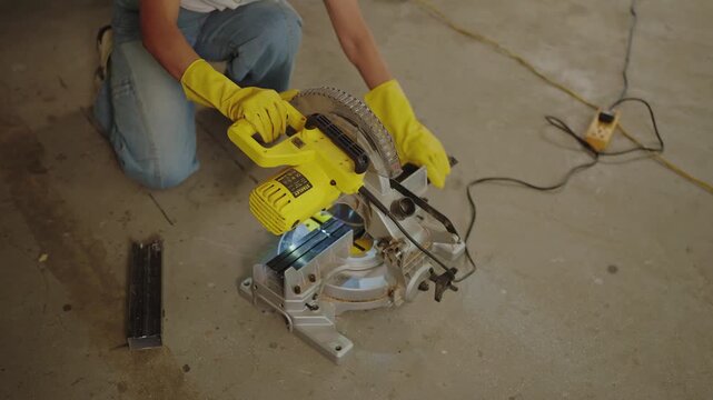 Overhead close up of gloved hands operating yellow miter saw cutting metal bracket with bright blue spark glow on concrete floor
