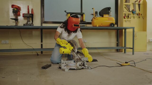 Young woman in white shirt and blue jeans kneeling to operate yellow miter saw cutting metal bracket in spacious industrial workshop