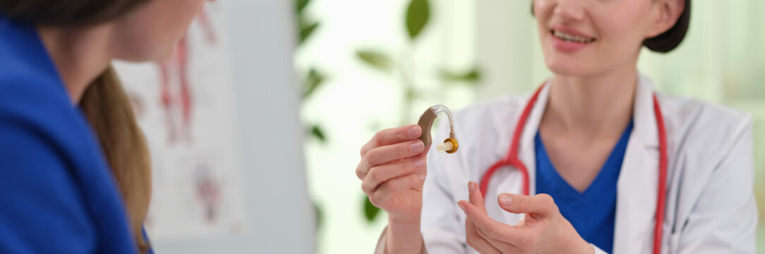 Woman doctor shows hearing aid during audiology consultation in clinic. Smiling specialist presents small device explaining to patient sound support